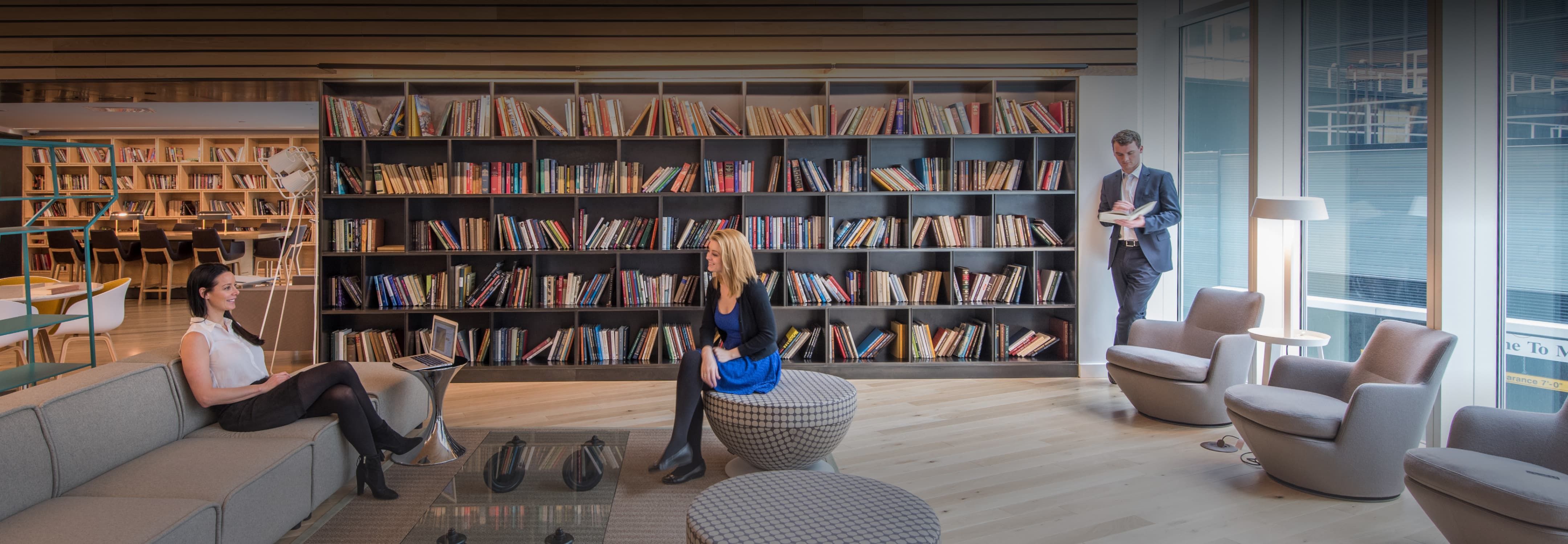 Modern library space with patrons, large bookshelves, comfortable seating, and large windows.